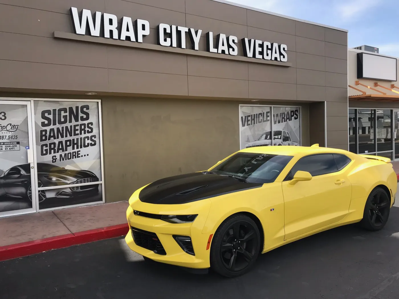 Yellow coupe with a matte black hood and roof stripe, parked at the storefront.