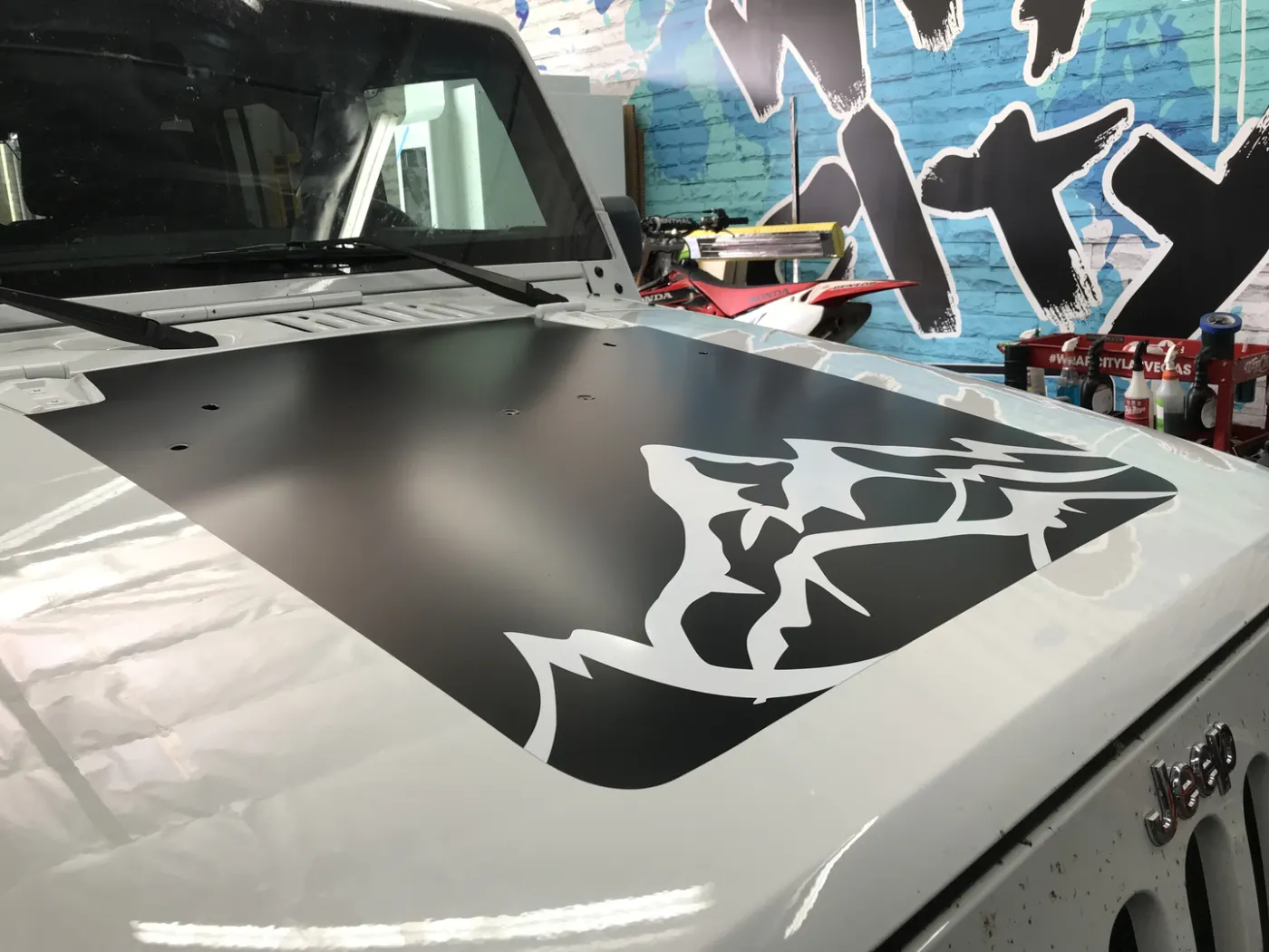 Matte black hood wrap with a cut graphic knocked out of it, over a white coupe.