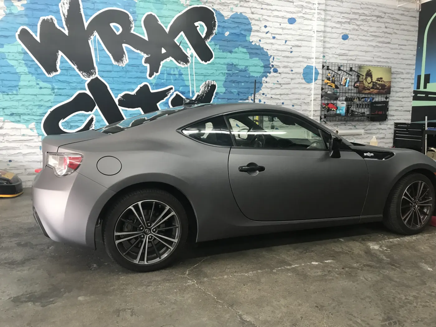 Sports coupe in a matte grey colour change, shot against the shop's painted logo wall.