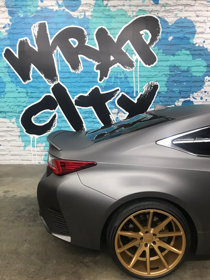 Rear three-quarter of a matte colour change with gold wheels, against the logo wall.